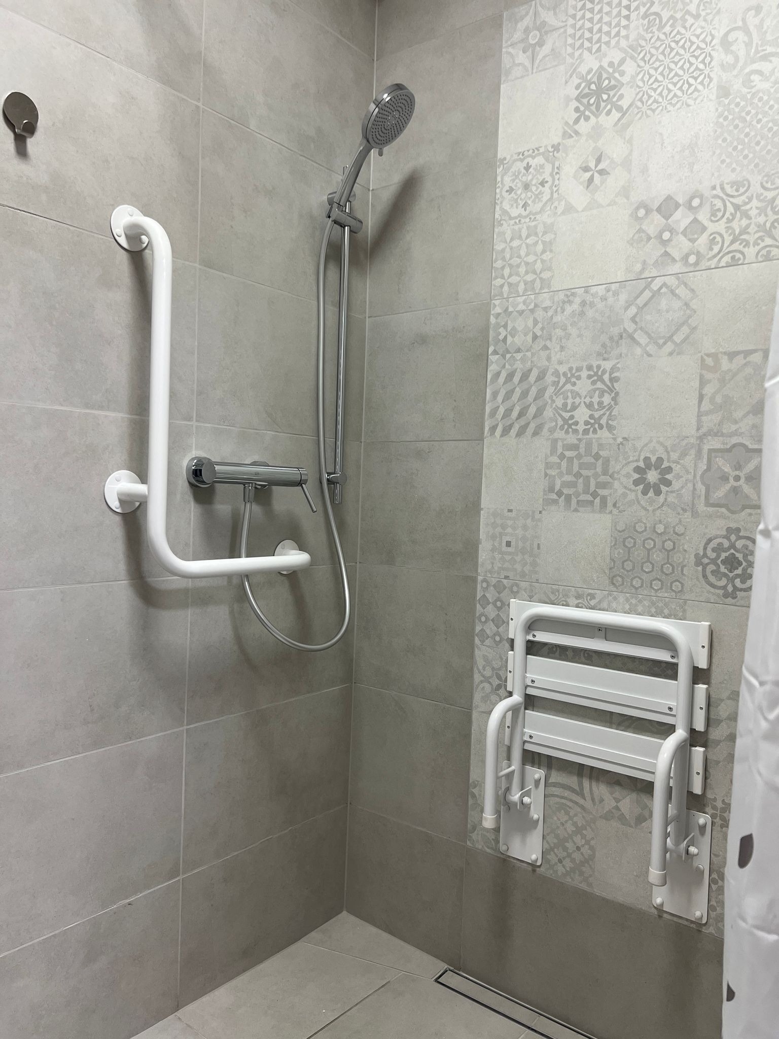 Wnętrze łazienki z zamontowanym chromowanym zestawem prysznicowym, białą poręczą dla niepełnosprawnych oraz składanym siedziskiem prysznicowym na ścianie z dekoracyjnymi płytkami.