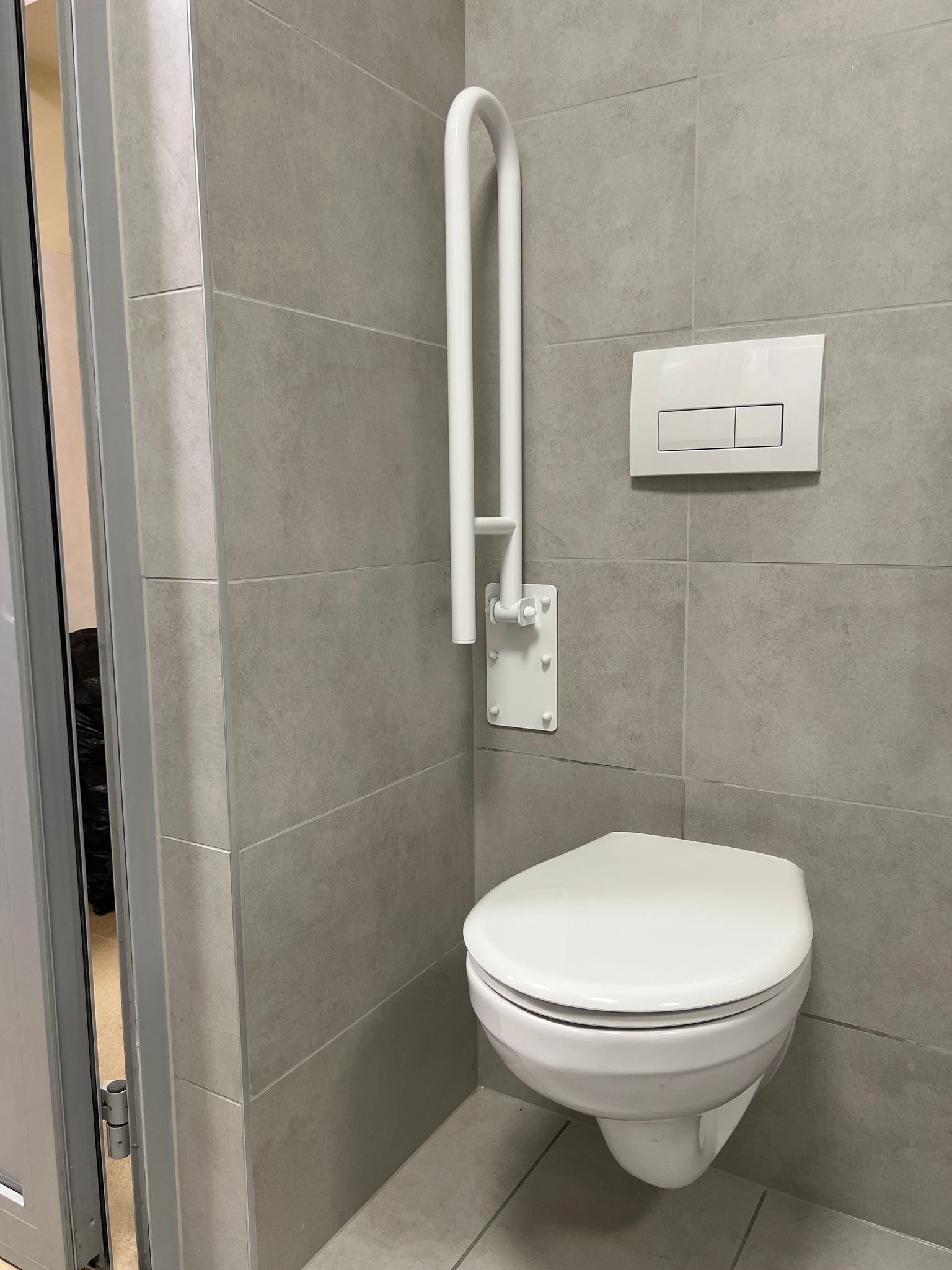 Nowoczesna toaleta podwieszana z białym przyciskiem spłukującym i uchwytem dla niepełnosprawnych, zamontowana na szarych płytkach ceramicznych w łazience.