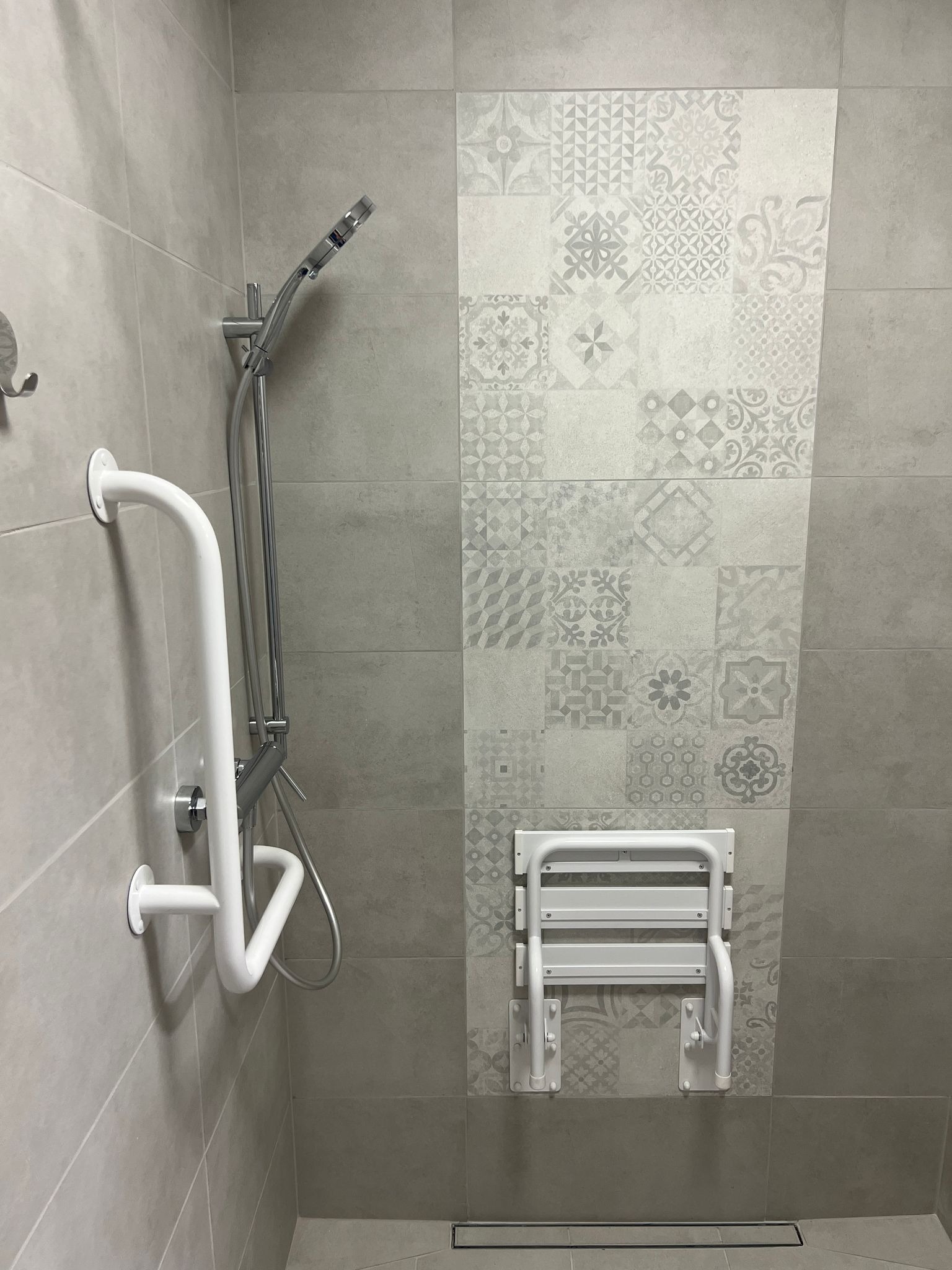 Wykończona łazienka z szarymi płytkami, dekoracyjnym pasem mozaiki, uchwytem dla niepełnosprawnych przy natrysku oraz składanym siedziskiem prysznicowym.