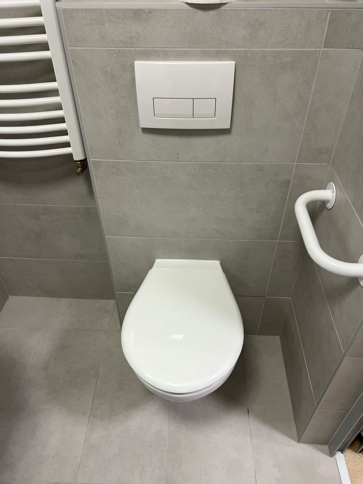 Nowoczesna toaleta z podwieszaną miską, przyciskiem spłukującym w kolorze białym, grzejnikiem drabinkowym i uchwytem dla niepełnosprawnych na szarej płytkowej ścianie.