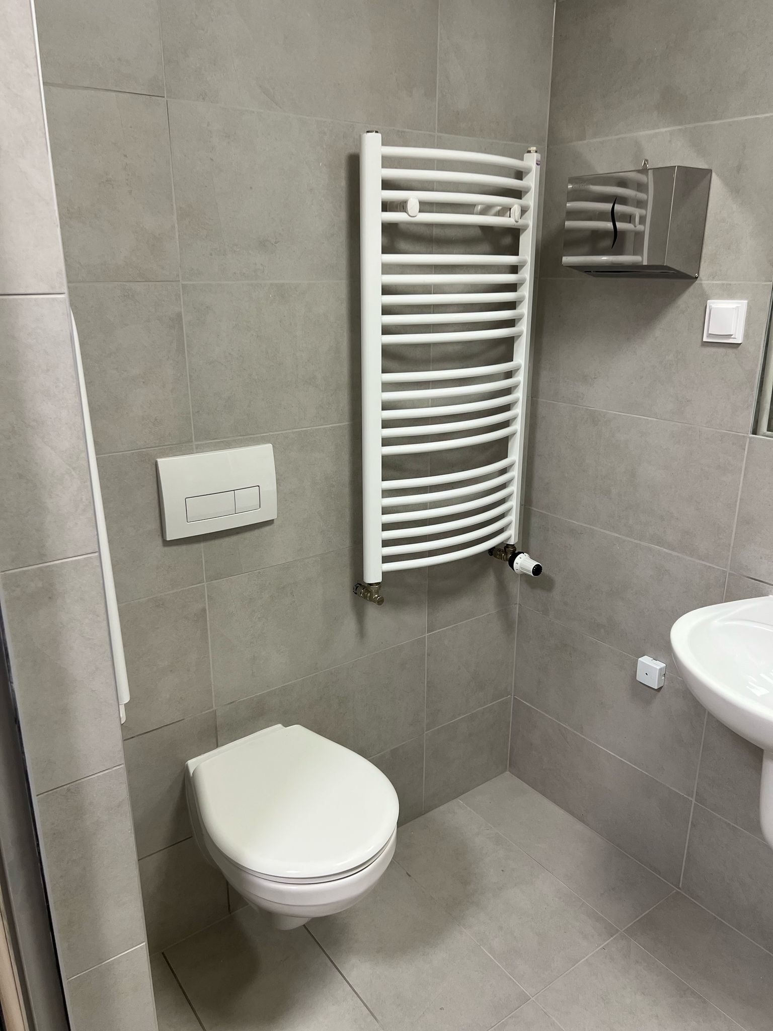 Nowoczesna łazienka z szarymi płytkami, wiszącą toaletą, białą drabinką grzejnikową z termostatem, podajnikiem na ręczniki papierowe i umywalką.