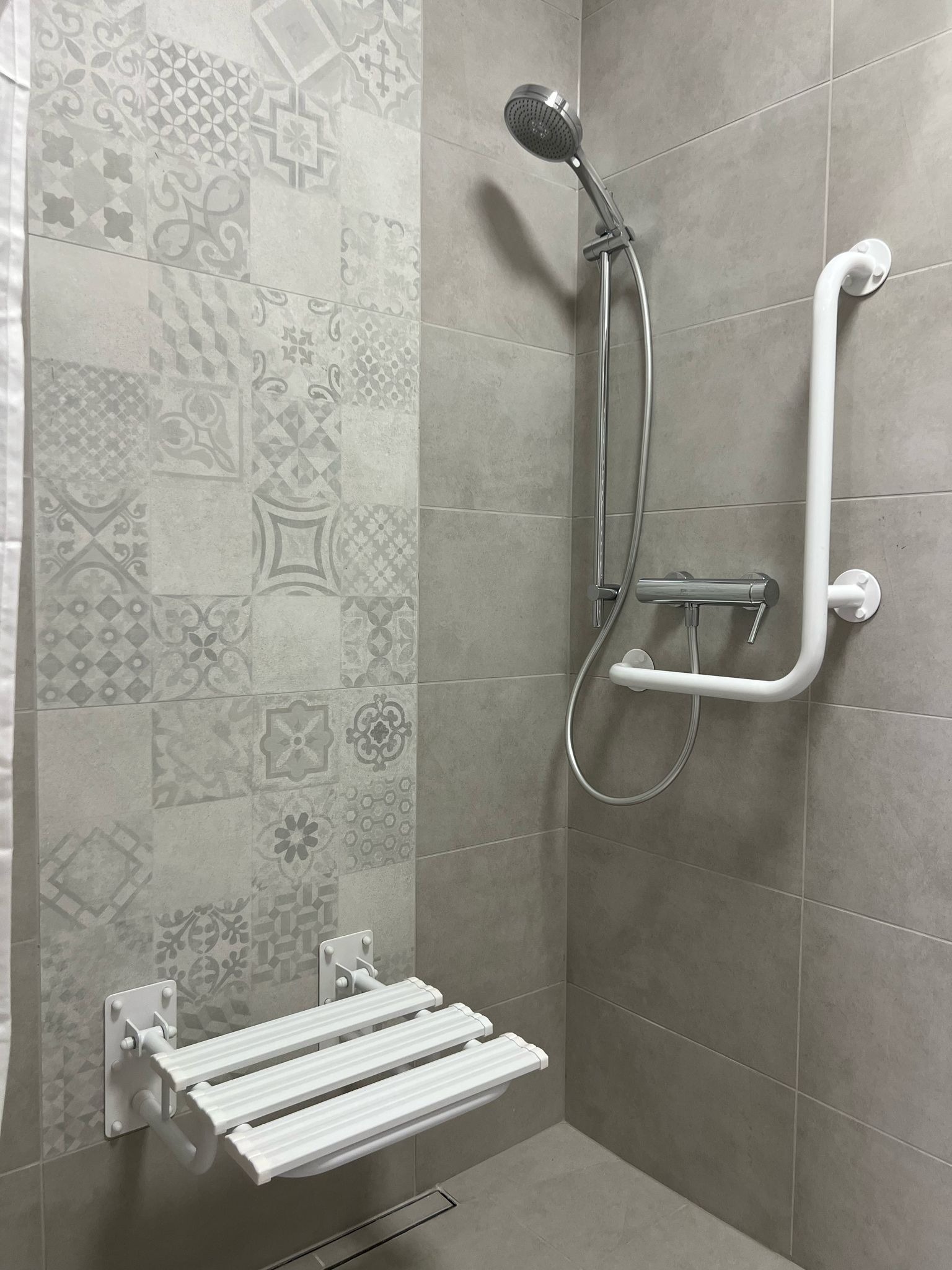 Nowoczesna kabina prysznicowa z siedziskiem dla osób z ograniczoną mobilnością i uchwytem, wykończona szarymi płytkami z dekoracyjnym wzorem.