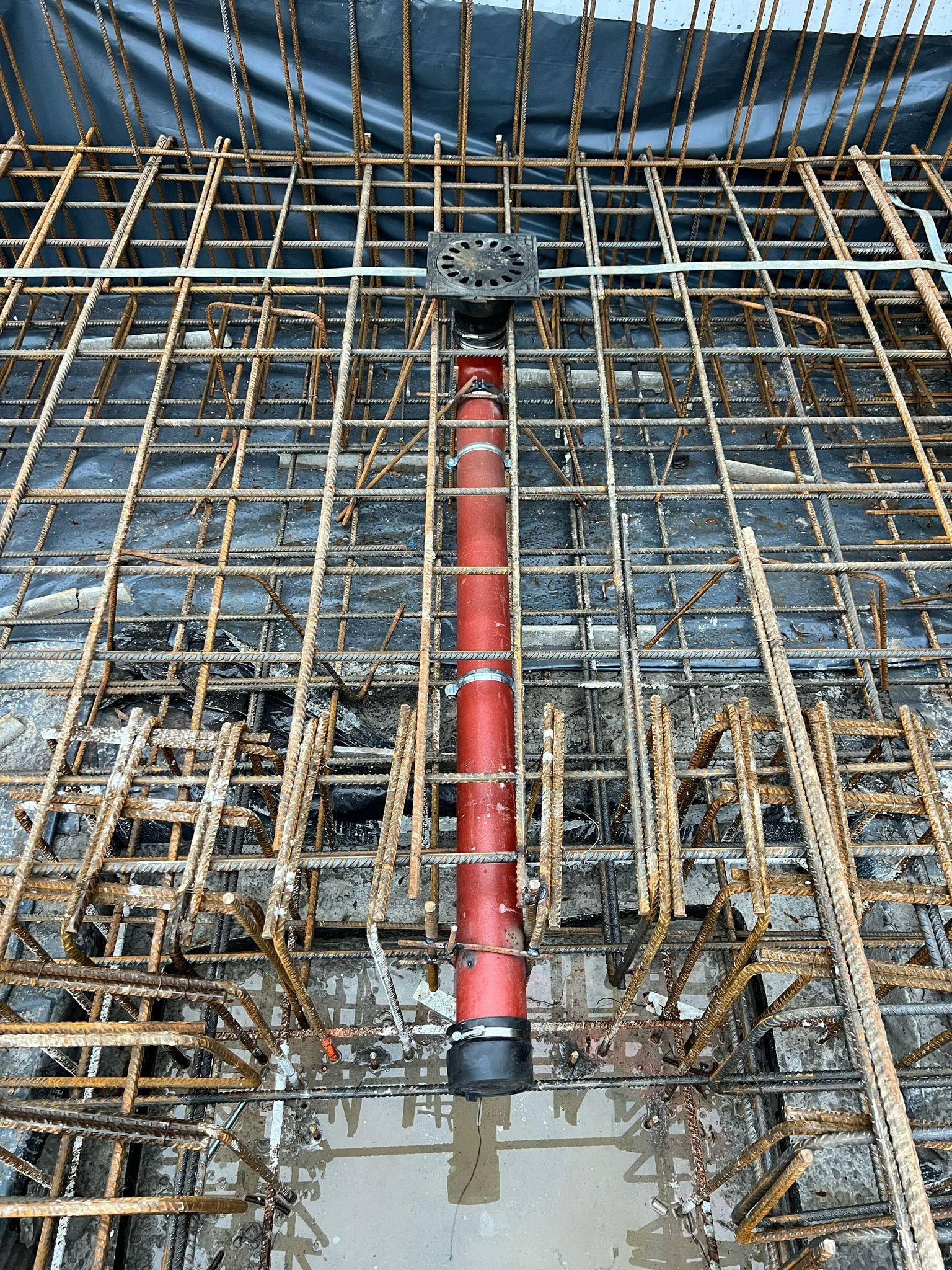 Widok z góry na czerwoną rurę kanalizacyjną z metalowymi obejmami, przechodzącą przez zbrojenie pod wylewkę betonową, z widocznym odpływem podłogowym i czarną folią w tle.