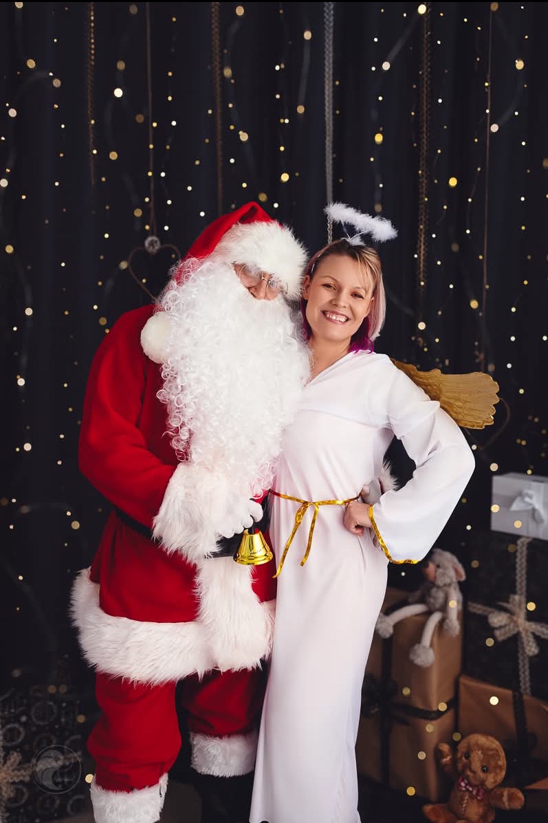 Uśmiechnięta kobieta w stroju anioła pozuje z Mikołajem trzymającym dzwonek na tle prezentów i ciemnej ściany z lampkami. Dekoracja świąteczna, impreza.