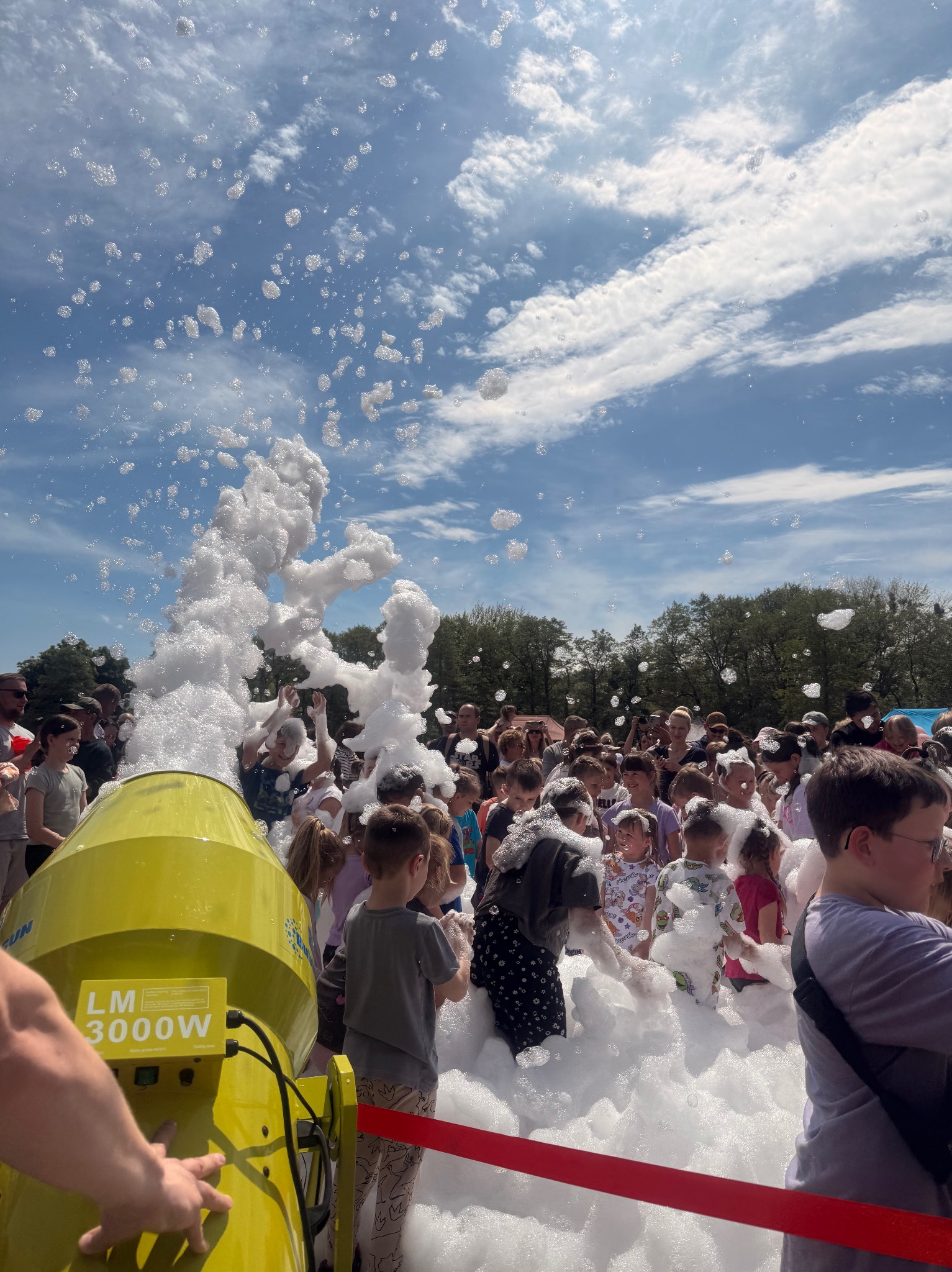 Żółta maszyna do piany LM 3000W otoczona bawiącymi się dziećmi, z pianą unoszącą się w powietrzu na tle błękitnego nieba z chmurami. Impreza plenerowa.