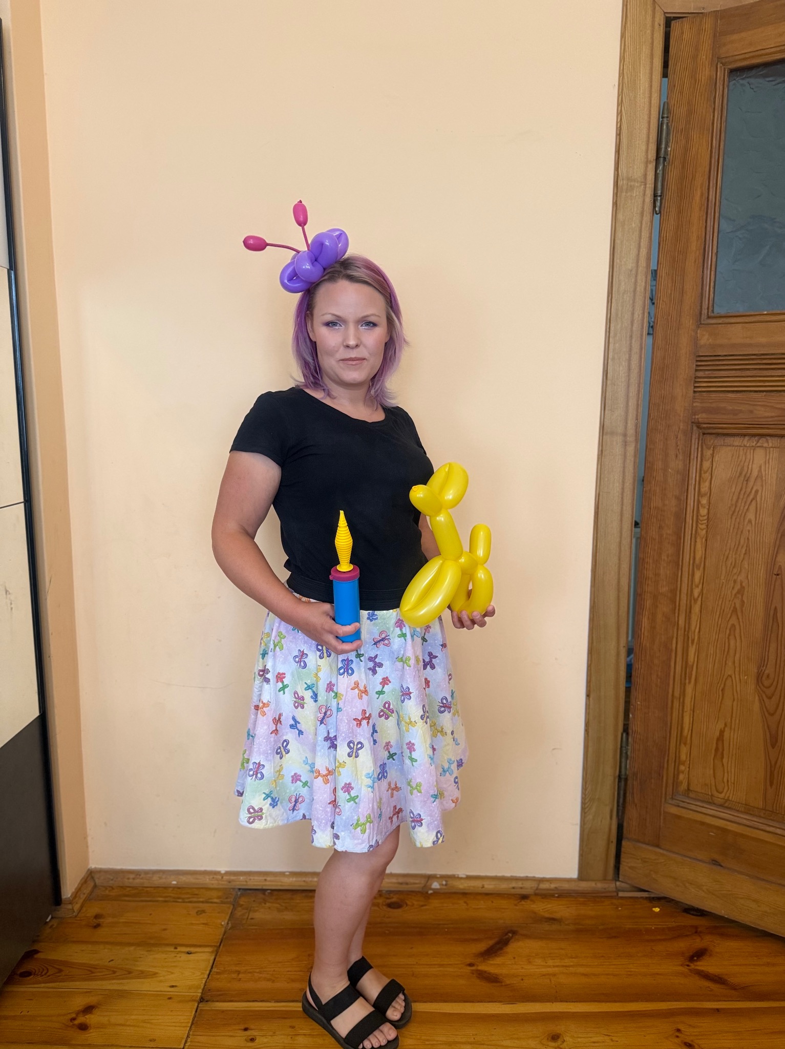 Animator w czarnej koszulce i spódnicy w balony trzyma pompkę i balon w kształcie psa. Na głowie ma ozdobę z balonów. W tle ściana i drewniane drzwi.