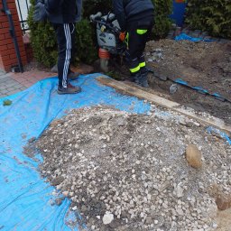 Daniel Gawęcki hydraulik kompleksowe usługi remontowo budowlane - Prace ziemne: zagęszczarka wibrująca ubija grunt pod przyszłą instalację, widoczny stos kruszywa na niebieskiej plandece, dwóch pracowników w odzieży roboczej.