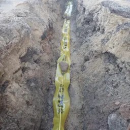 Wykop z ułożoną żółtą rurą ochronną z napisem PN-EN 12613, zabezpieczoną kamieniami, przygotowany do zasypania.