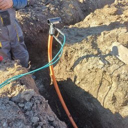 Daniel Gawęcki hydraulik kompleksowe usługi remontowo budowlane - Pomarańczowa rura gazowa lub wodociągowa w wykopie, podłączona do czarnej złączki, widoczne niebieskie przewody i fragment osoby w kombinezonie.