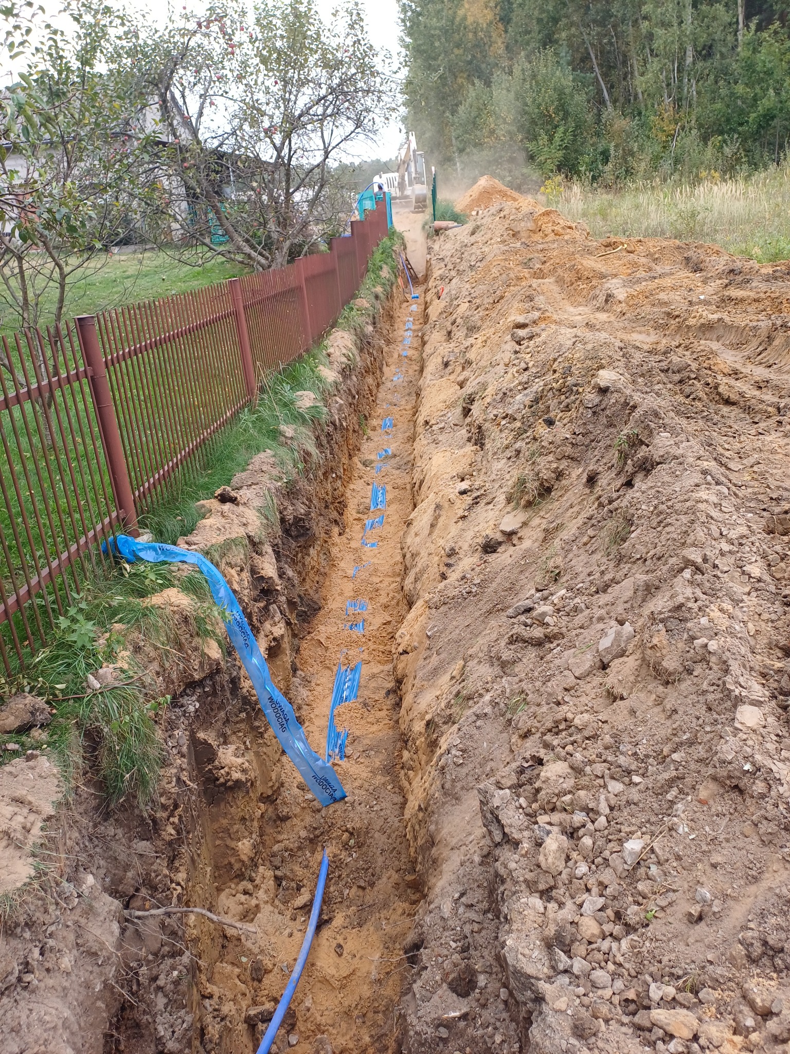 Wykop pod niebieski rurociąg w ziemi, zabezpieczony niebieską folią ostrzegawczą, widoczny fragment brązowego ogrodzenia i koparka w oddali.