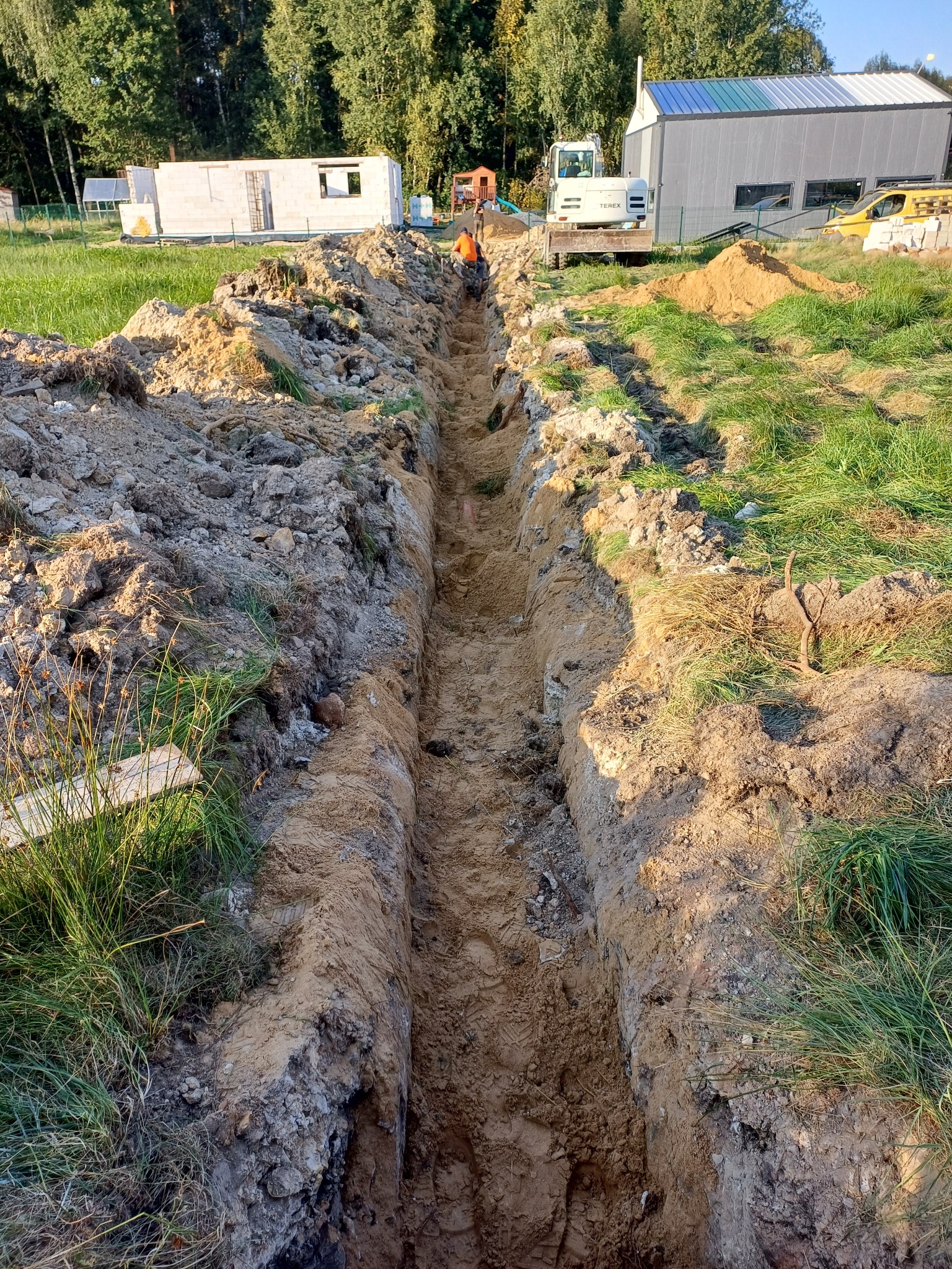 Wykopany rów w ziemi na trawiastym terenie, w oddali widoczny pracownik w pomarańczowym ubraniu i maszyny budowlane.