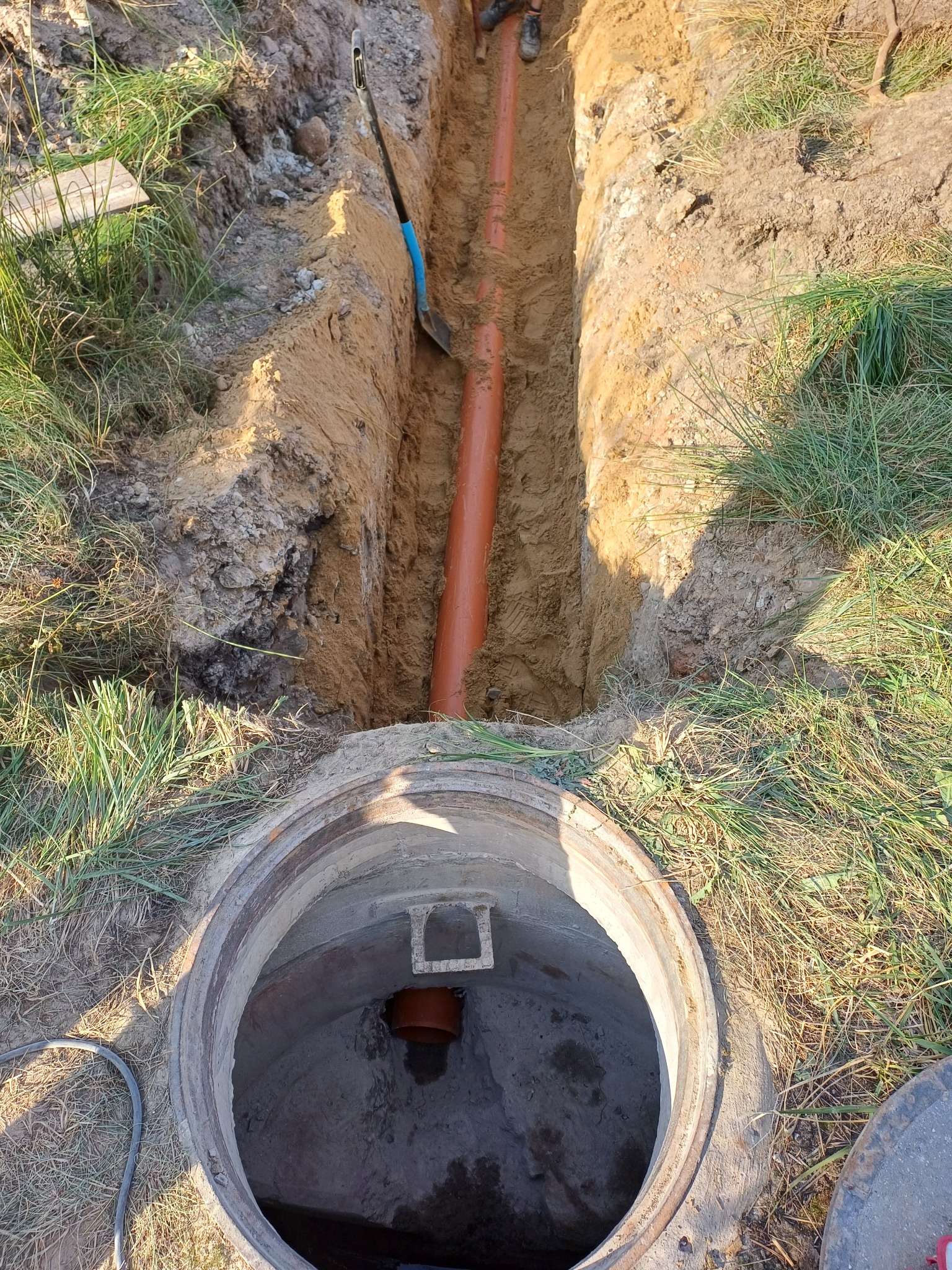 Wykonany wykop pod rurę kanalizacyjną, widoczny fragment rury w wykopie oraz studzienka kanalizacyjna z fragmentem rury wewnątrz, łopata wbita w ziemię obok wykopu.