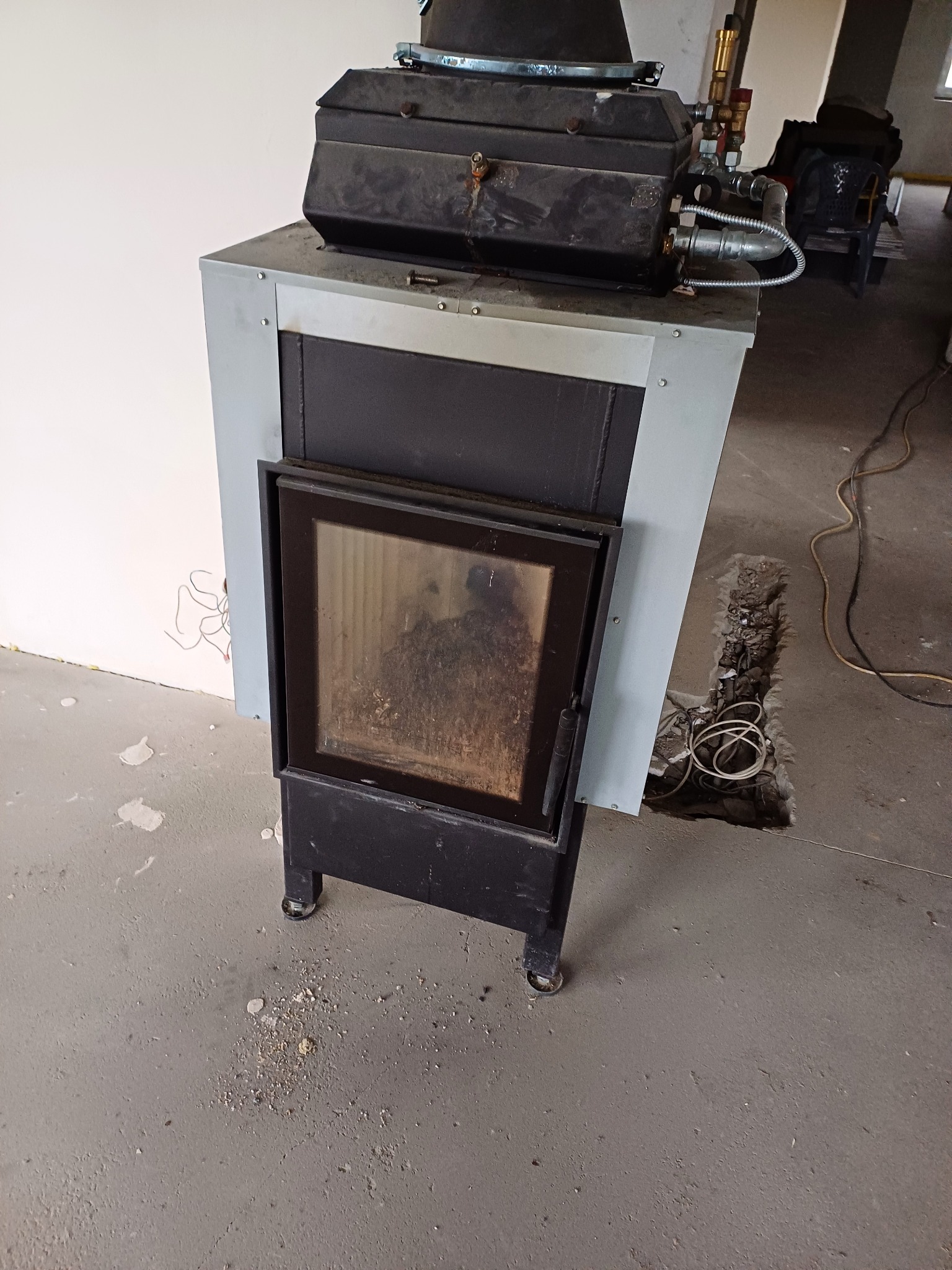 Stary piec gazowy z otwartymi drzwiczkami, widoczne przyłącza gazowe i wykop w podłodze, instalacja w trakcie prac remontowych.
