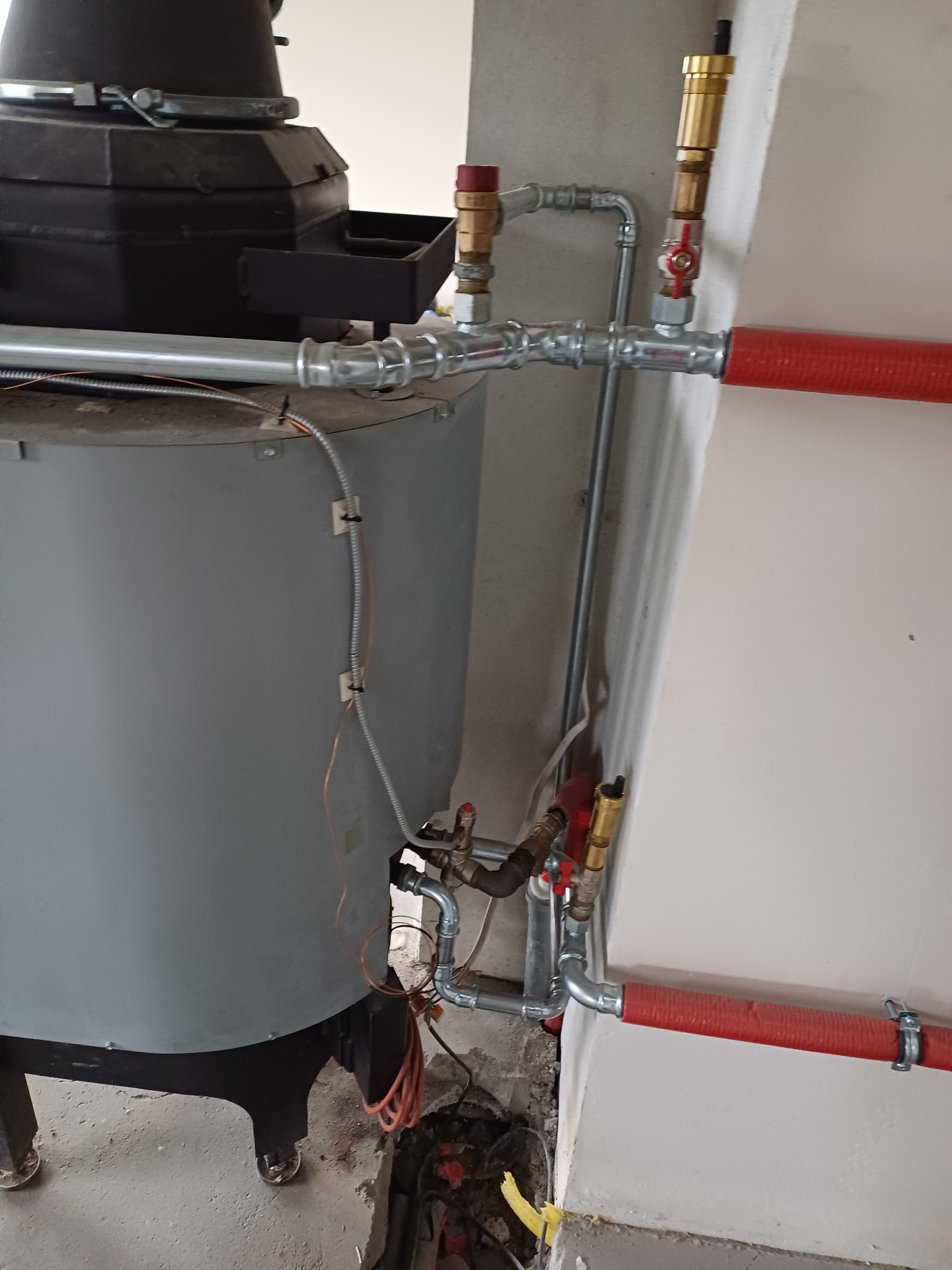Szary piec z czarnym podajnikiem, rury instalacji centralnego ogrzewania z zaworami i czerwoną izolacją, widoczne okablowanie i fragment podłogi w trakcie instalacji.