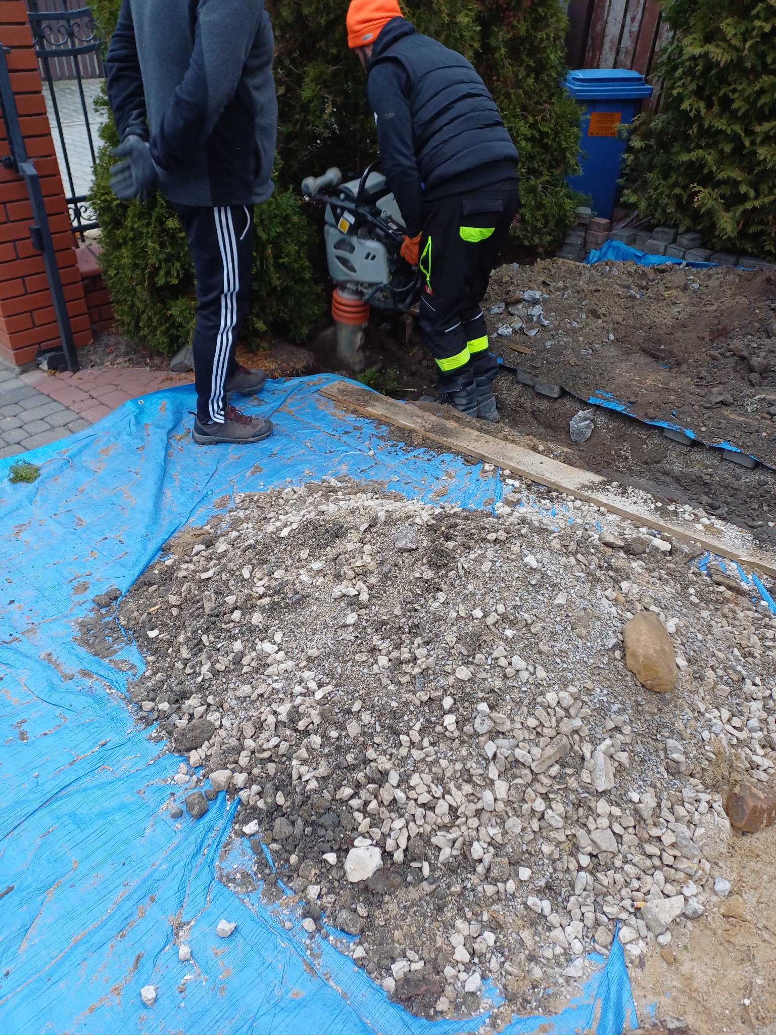 Prace ziemne: zagęszczarka wibrująca ubija grunt pod przyszłą instalację, widoczny stos kruszywa na niebieskiej plandece, dwóch pracowników w odzieży roboczej.