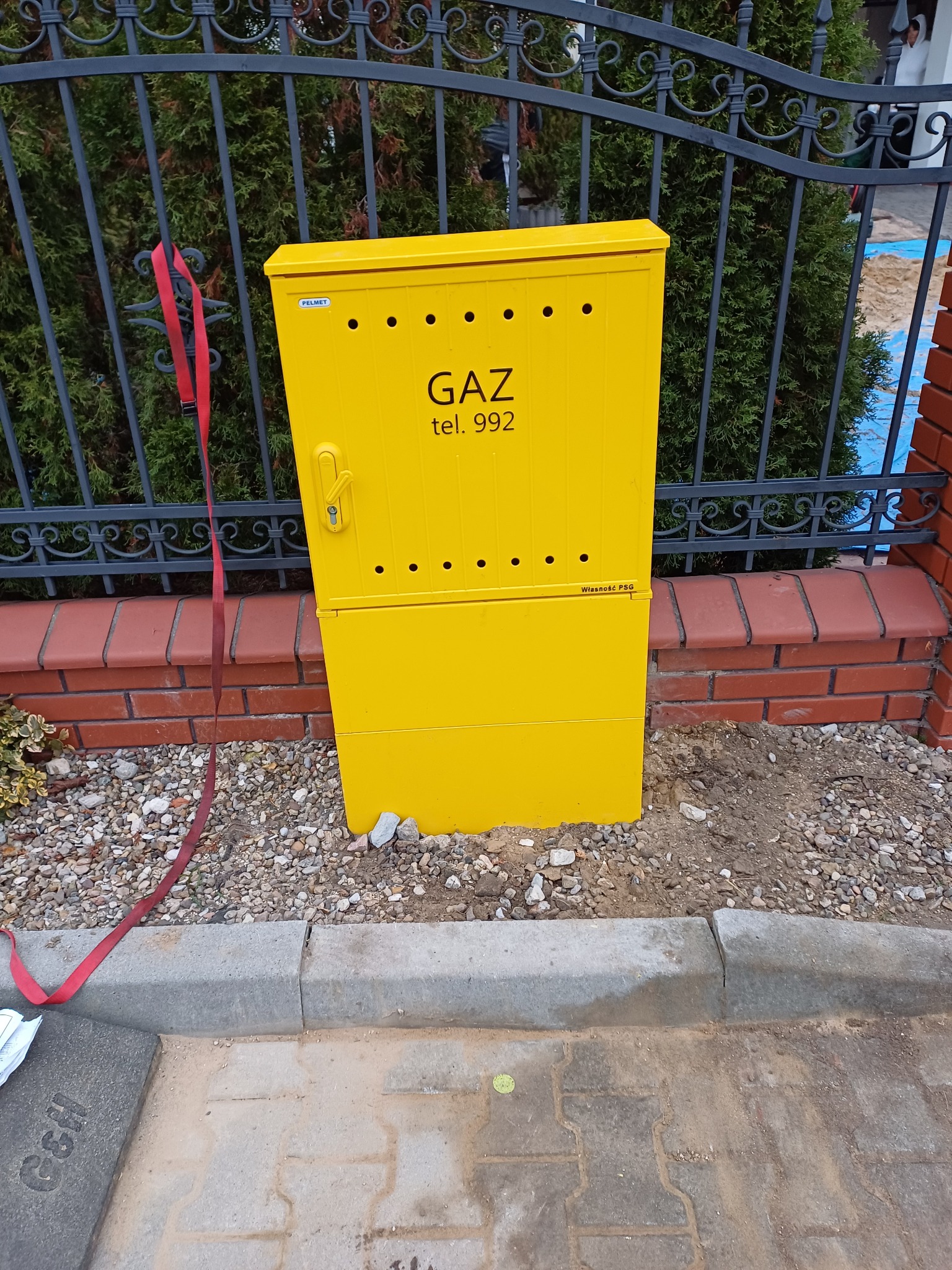 Żółta skrzynka gazowa z napisem 'GAZ tel. 992' umieszczona przy ogrodzeniu z metalowymi prętami i ceglanym murkiem, widoczny czerwony pas zwisający z ogrodzenia.