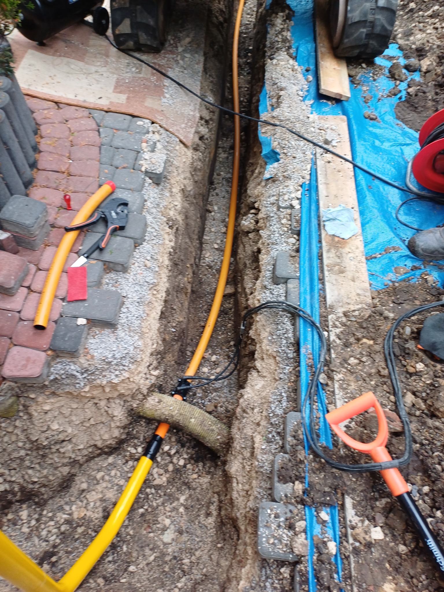 Wykop z ułożonymi rurami gazowymi i niebieskimi rurami osłonowymi, widoczne narzędzia i fragment kostki brukowej. Wykop zabezpieczony folią.