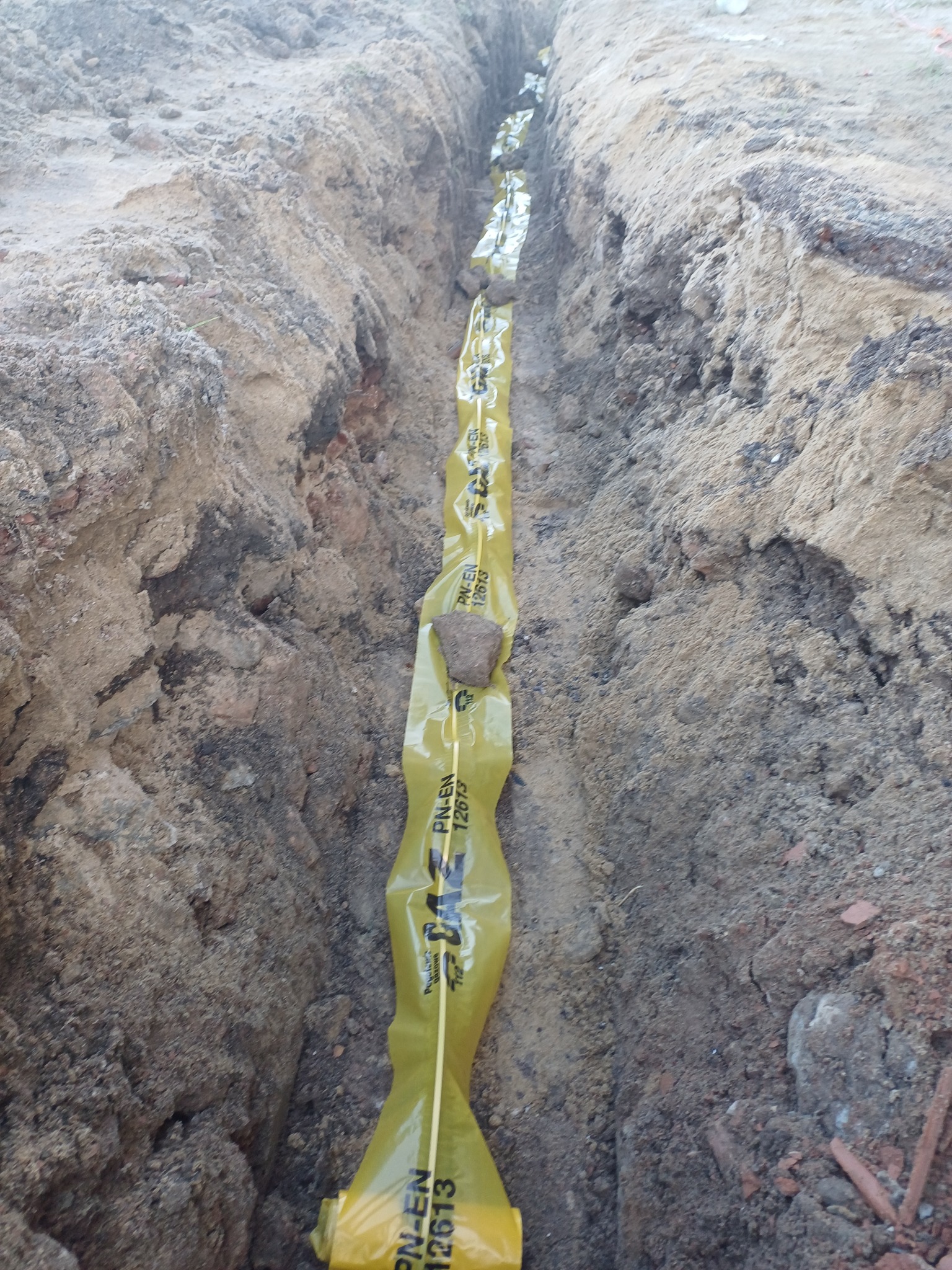 Wykop z ułożoną żółtą rurą ochronną z napisem PN-EN 12613, zabezpieczoną kamieniami, przygotowany do zasypania.