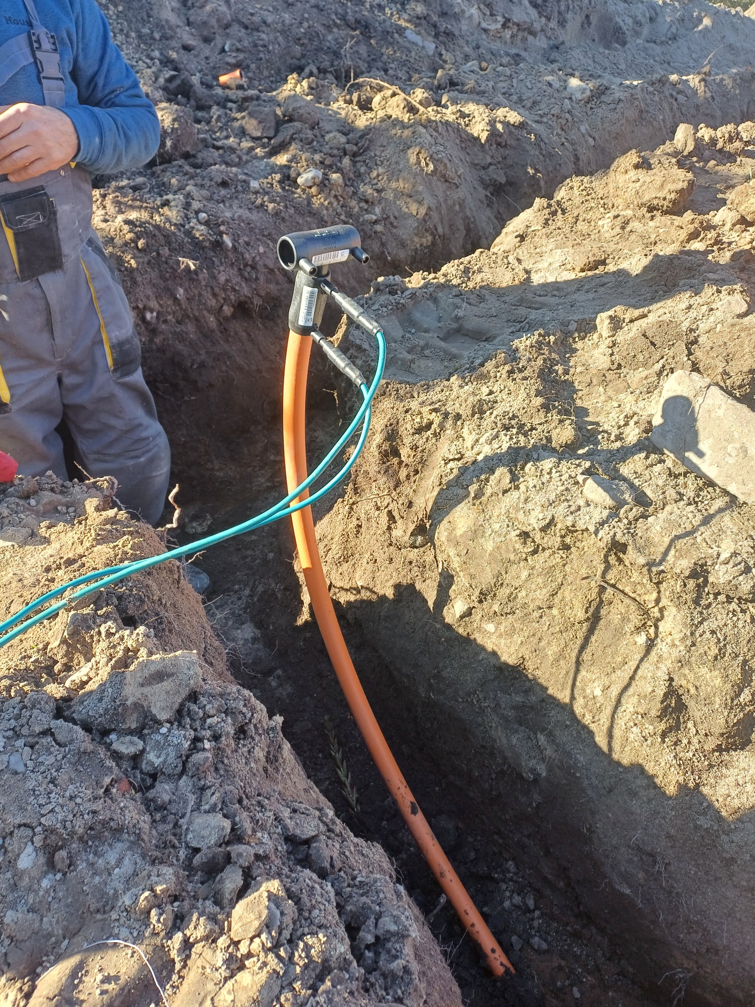 Pomarańczowa rura gazowa lub wodociągowa w wykopie, podłączona do czarnej złączki, widoczne niebieskie przewody i fragment osoby w kombinezonie.