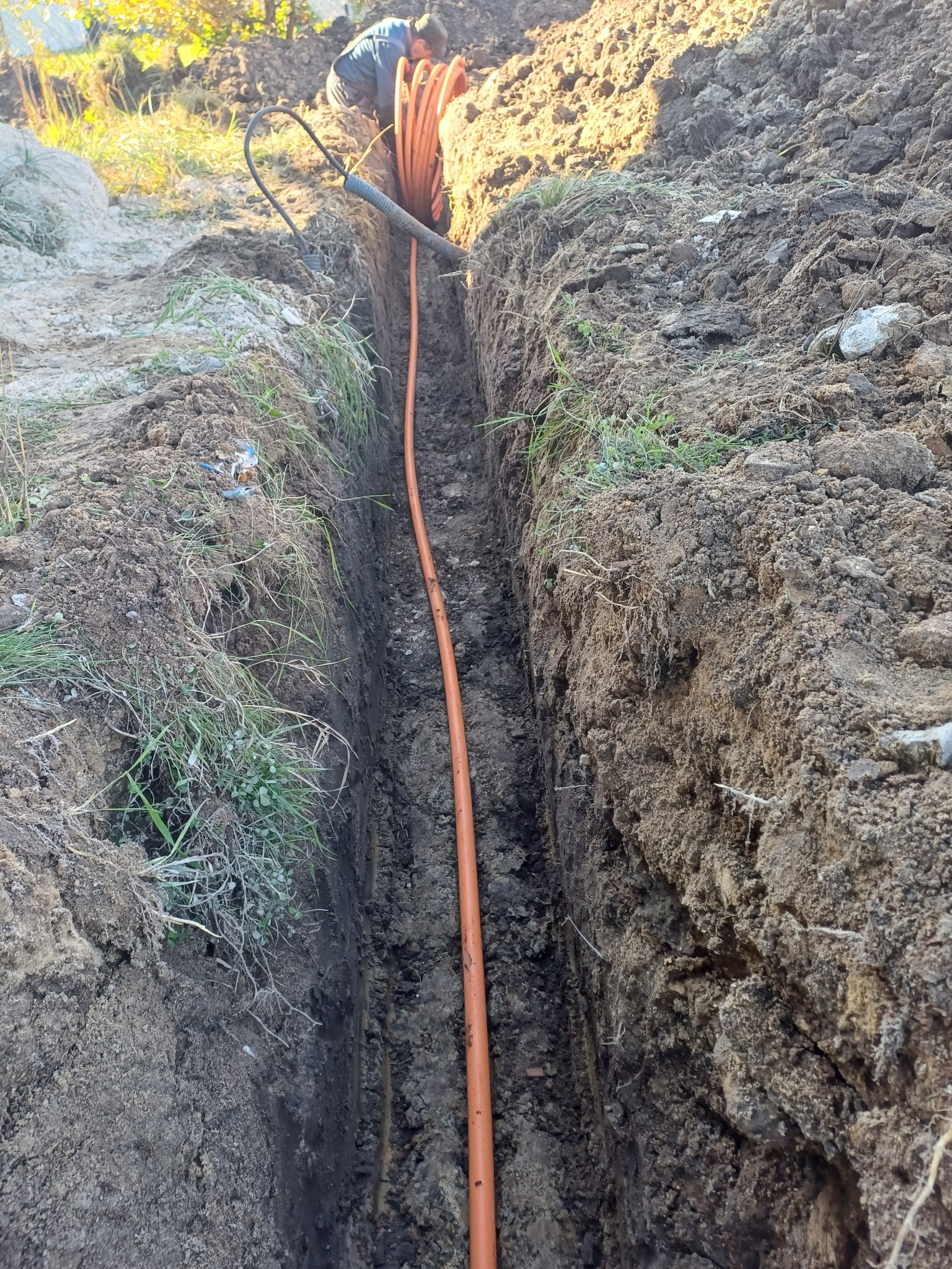 Pomarańczowa rura ułożona w wykopie ziemnym, widoczny fragment osoby pracującej przy instalacji.