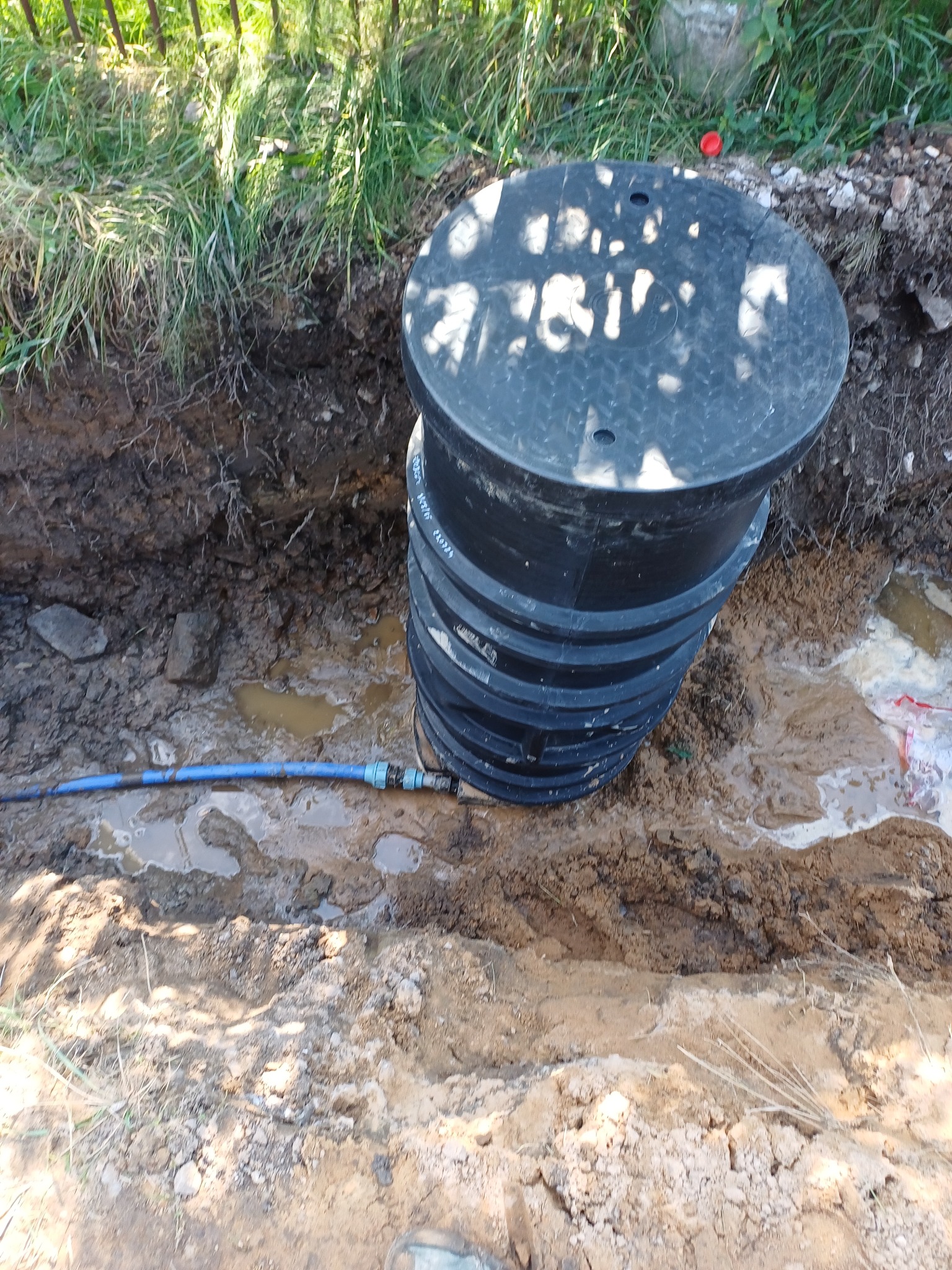 Czarny, plastikowy zbiornik na szambo lub deszczówkę w wykopie, podłączony do niebieskiej rury wodociągowej z widocznymi złączkami, na tle mokrej ziemi i trawy.