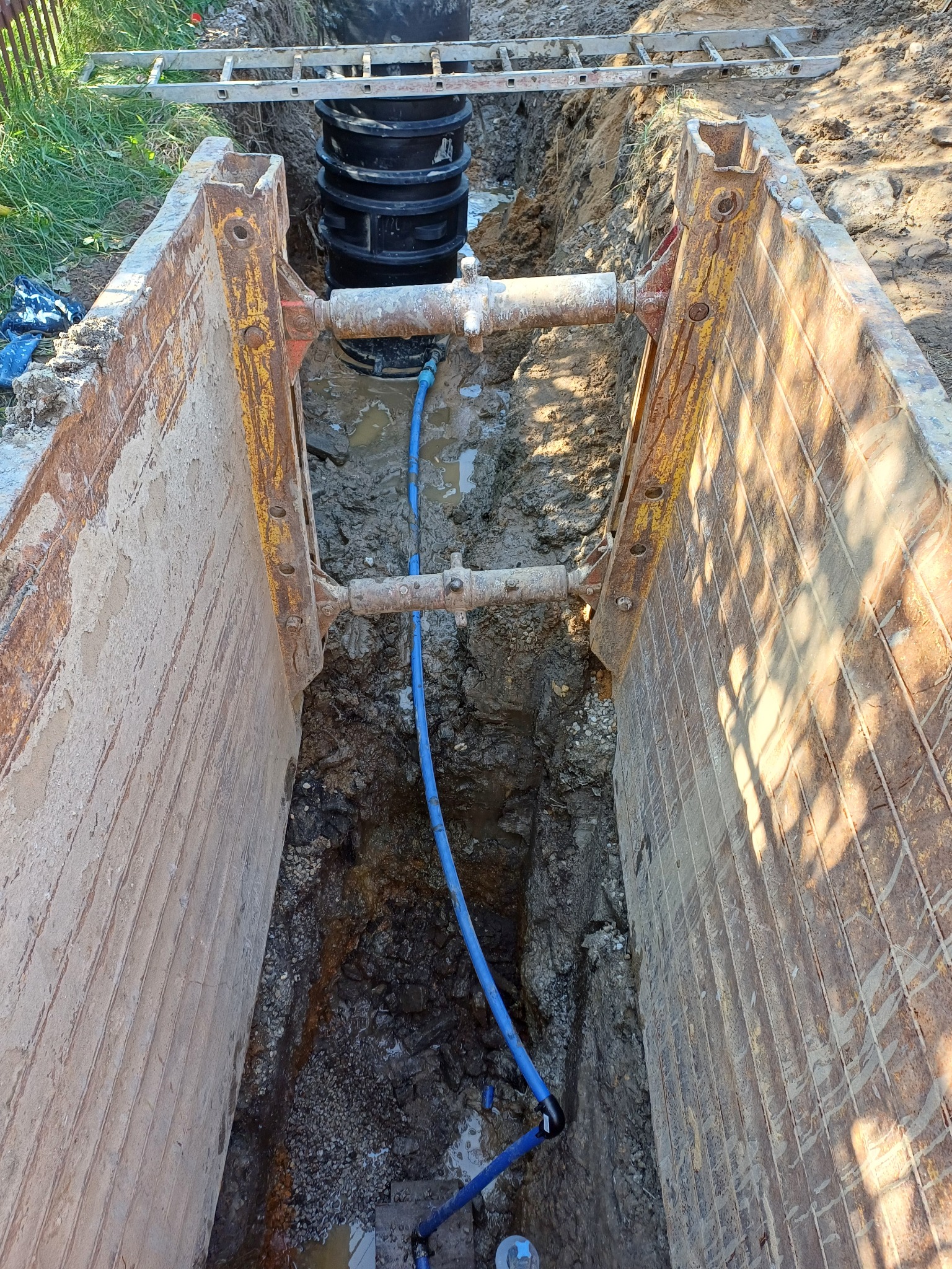 Głęboki wykop zabezpieczony stalowymi szalunkami, z widoczną rurą wodociągową z tworzywa sztucznego i czarnym zbiornikiem na dnie wykopu, drabina na górze.