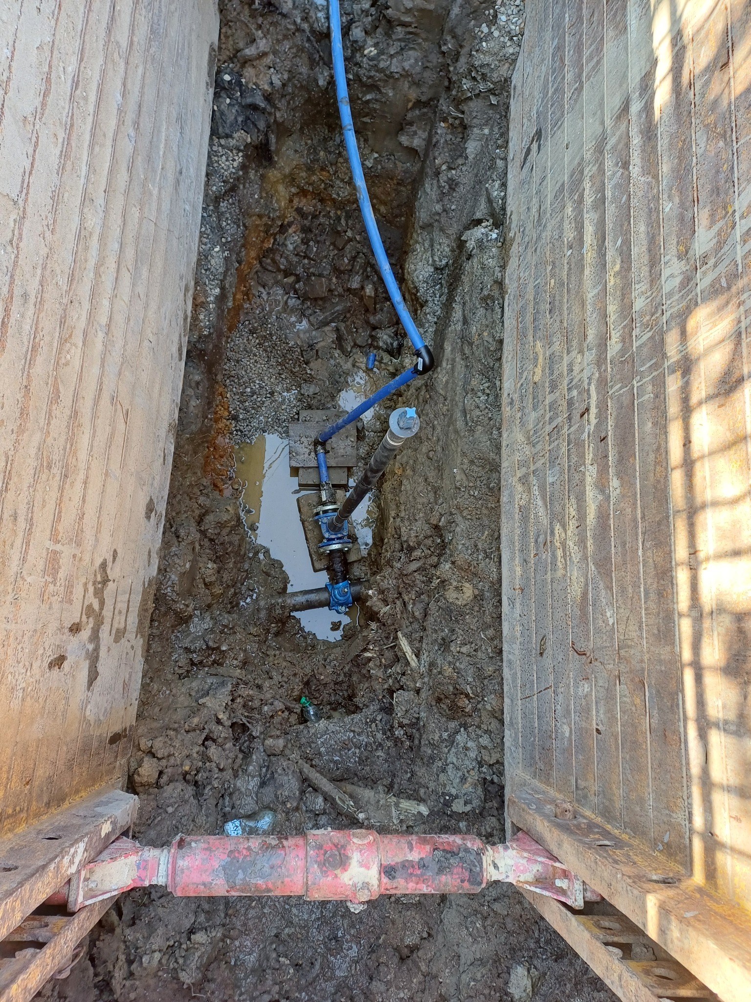 Wykop zabezpieczony szalunkiem z widocznym fragmentem nowej instalacji wodociągowej z rur PE, częściowo zanurzonej w wodzie i błocie.