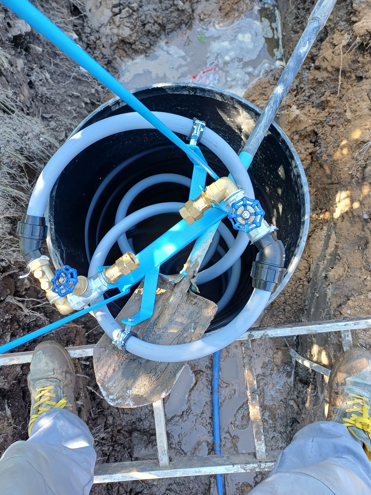 Wnętrze czarnego zbiornika z szarymi rurami, zaworami z niebieskimi pokrętłami i niebieskim wspornikiem, widoczne z góry, z elementami szpadla i butami pracownika na metalowej drabince.