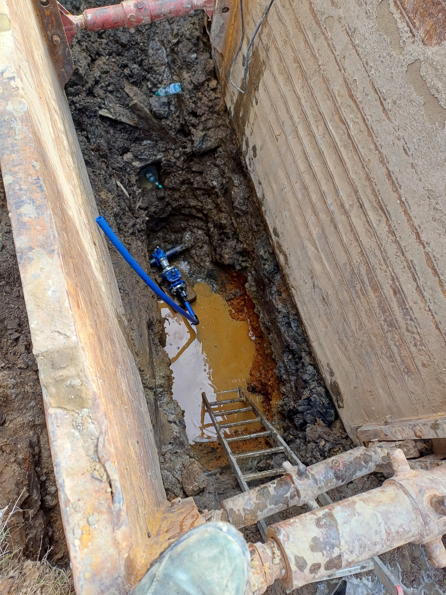 Wykop zabezpieczony szalunkiem stalowym z widoczną instalacją wodociągową z niebieską rurą i zaworem oraz drabinką w brudnej, zalanej wodą wykopie.