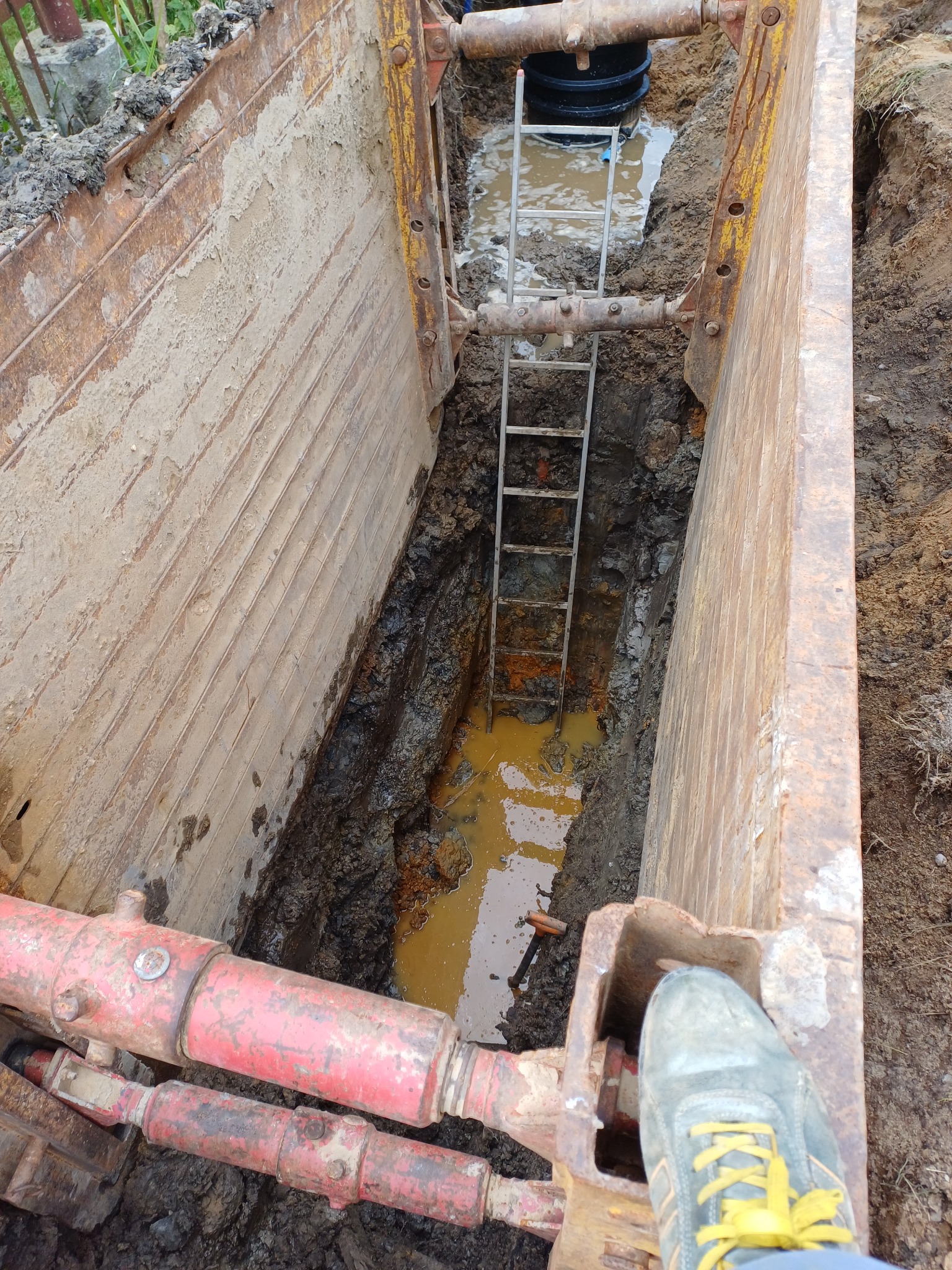 Wykonany wykop zabezpieczony stalowymi szalunkami z widoczną drabiną i fragmentem rurociągu w błotnistej wodzie, hydrauliczne rozpory w kolorze czerwonym.