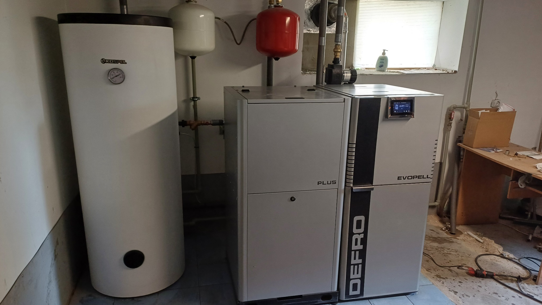 Biały piec Defro Evopell Plus z zbiornikiem Kostrzewa, instalacja CO w kotłowni. Widoczne rury, zawory i czujnik temperatury. Niedokończona posadzka.