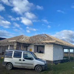 Budowa domu jednorodzinnego z widoczną konstrukcją dachu, mury z białych bloczków, srebrny samochód dostawczy na pierwszym planie, błękitne niebo z chmurami w tle.