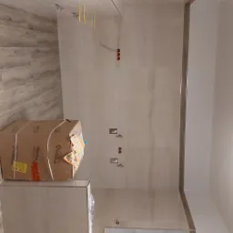 Łazienka w trakcie wykańczania z beżowymi płytkami imitującymi marmur, widoczne przygotowane otwory na armaturę, karton z zapakowanymi elementami wyposażenia i okno.