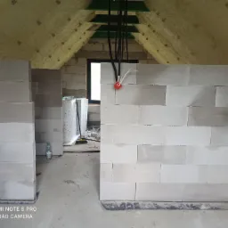 Wnętrze poddasza w trakcie budowy ścian działowych z bloczków, widoczne ocieplenie dachu pianą, instalacja elektryczna i materiały budowlane w tle.