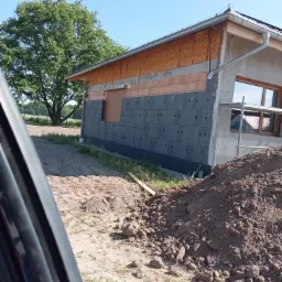 Budynek w trakcie ocieplania, widoczne szare płyty izolacyjne z czarnymi kołkami mocującymi, częściowo zamontowane, fragment okna z rusztowaniem, stos ziemi w pierwszym planie.