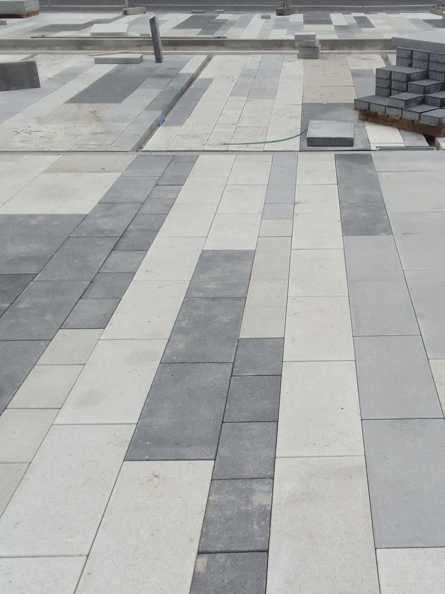 Fragment powierzchni z ułożoną, dwukolorową kostką w trakcie prac. Widoczne przygotowane elementy do dalszego układania i niezakończone fragmenty nawierzchni.
