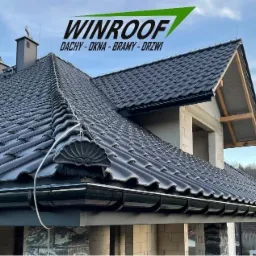 Nowy dom w trakcie budowy z ciemną dachówką ceramiczną i systemem rynnowym, widoczny logo firmy WINROOF na górze zdjęcia.