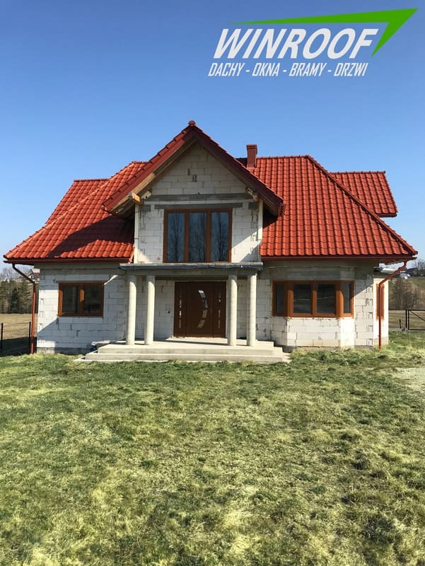 Dom w trakcie budowy z czerwoną dachówką, brązowymi oknami i drzwiami, widoczny na tle zielonej trawy i błękitnego nieba, z logo firmy WINROOF w górnej części zdjęcia.