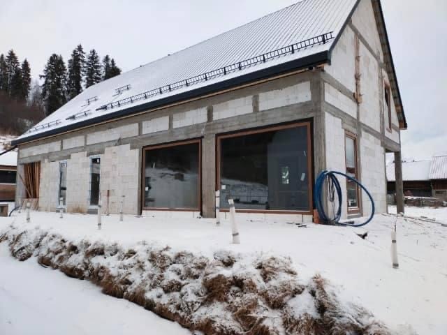 Budynek w trakcie budowy w zimowej scenerii, widoczne duże przeszklenia z brązowymi ramami, ściany z białych bloczków, częściowo ośnieżony dach z blachy, niebieski wąż leżący na śniegu przy ścianie.