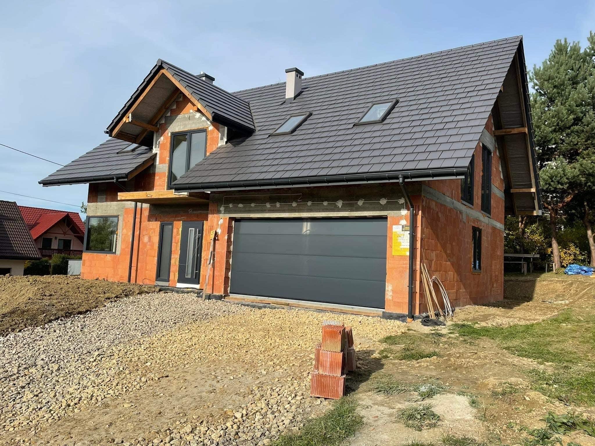 Niedokończony dom jednorodzinny z szarą dachówką i zamontowanymi oknami, w trakcie budowy, widoczne cegły i elementy konstrukcyjne, teren wokół domu częściowo zagospodarowany.