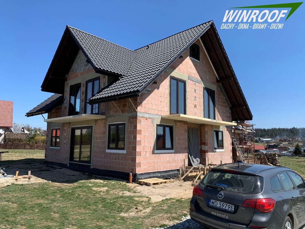 Dom w stanie surowym otwartym z zamontowanymi oknami i pokryciem dachowym, widoczny samochód osobowy z logo firmy WINROOF i numerem rejestracyjnym.