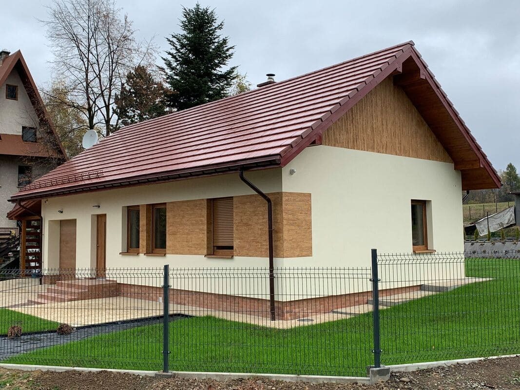 Dom jednorodzinny z czerwoną dachówką, kremową elewacją i drewnopodobnymi panelami dekoracyjnymi, otoczony zielonym trawnikiem i siatką ogrodzeniową.