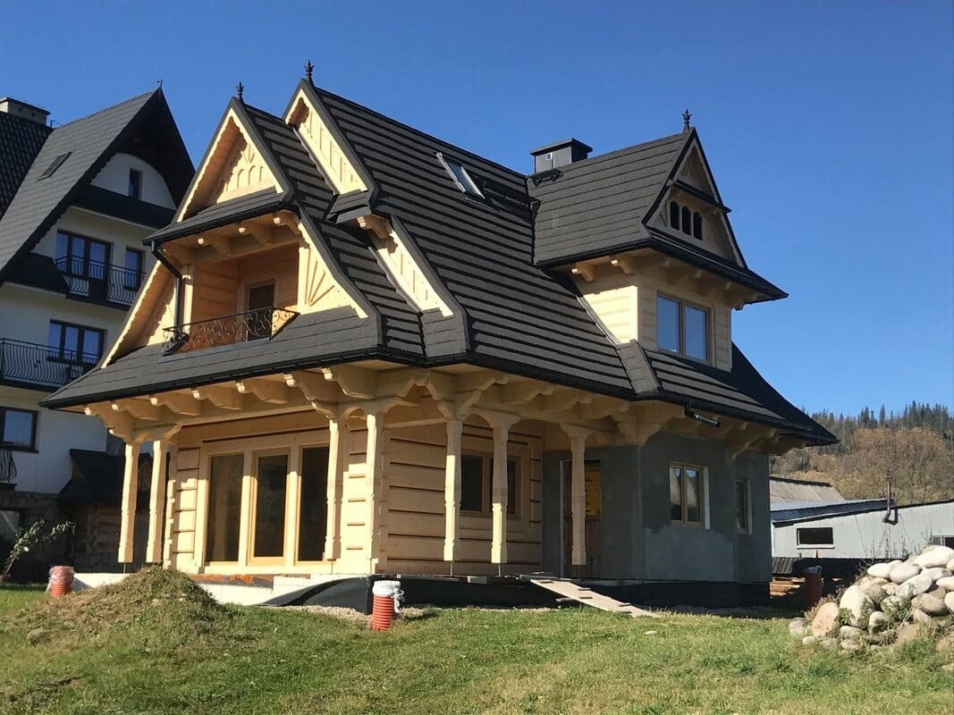 Surowy drewniany dom w stylu góralskim z ciemną dachówką, częściowo otynkowany, widoczny na tle błękitnego nieba i innych budynków.