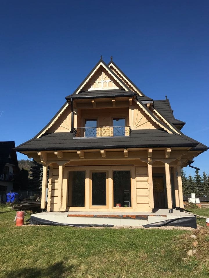 Drewniany dom w stylu góralskim z czarnym dachem, balkonem z ozdobną balustradą i dużymi oknami bez ram na parterze, w trakcie wykończenia.