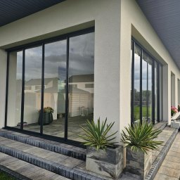 ALU-MAX ROBERT SZCZEPANIUK - Nowoczesny dom z dużymi, przesuwnymi oknami z czarnymi ramami, widoczne dwustopniowe schody z szarej kostki brukowej i rośliny w betonowych donicach, w tle odbicie ogrodu w szybach.