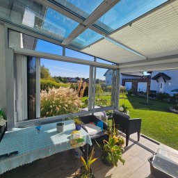 ALU-MAX ROBERT SZCZEPANIUK - Szklana zabudowa tarasu z aluminiową konstrukcją i roletami plisowanymi, widok na ogród z ozdobnymi trawami i domami w tle.