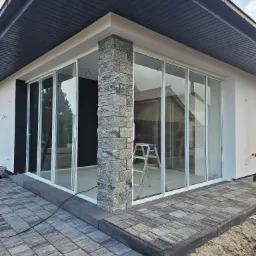 Nowoczesny dom z dużymi, przesuwnymi drzwiami szklanymi, wykończony kamienną kolumną i kostką brukową. W tle widoczna drabina.