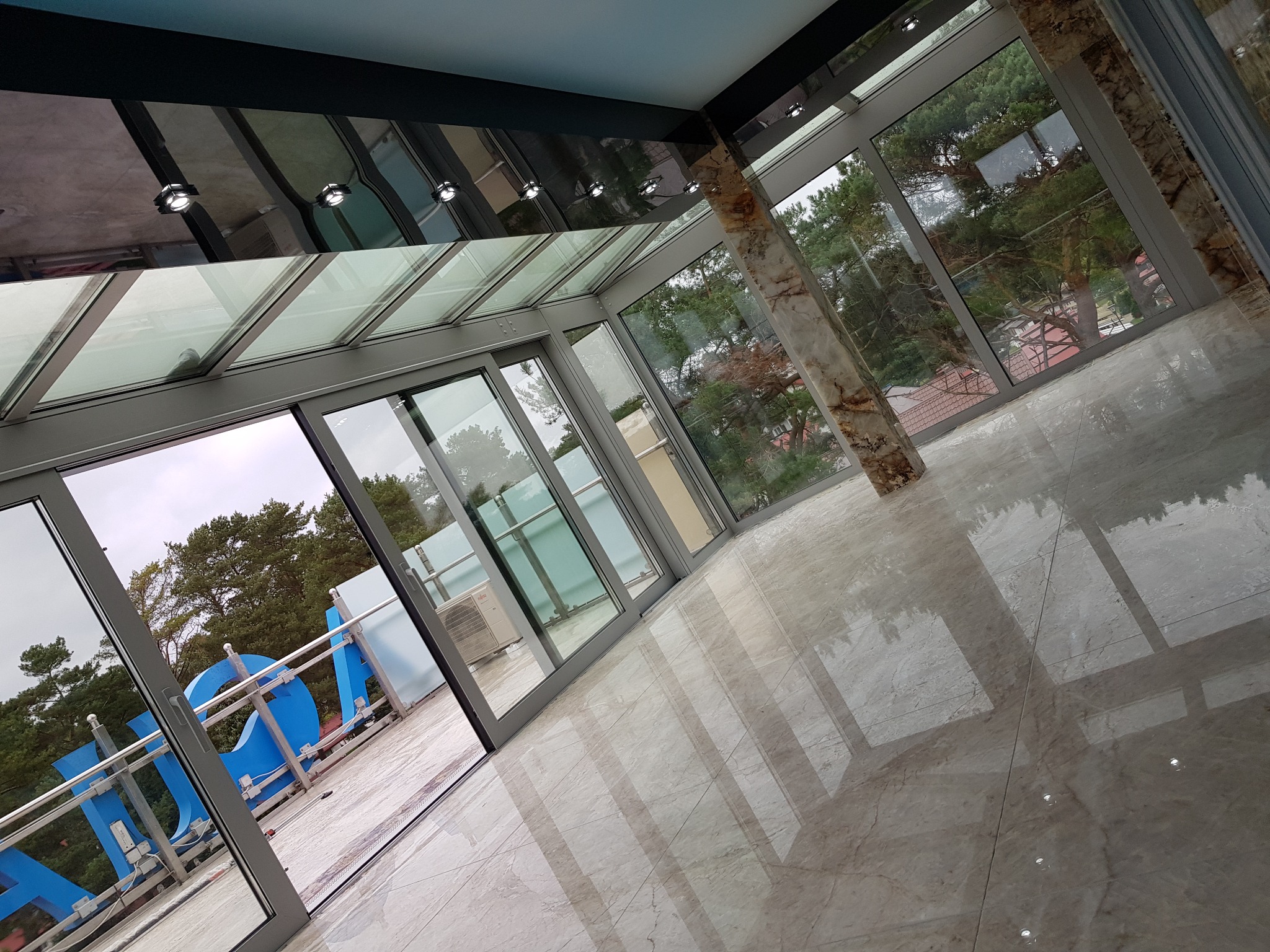 Nowoczesna oranżeria z przeszklonym dachem i przesuwnymi drzwiami, marmurowe filary i podłoga, widok na zewnątrz przez okna.