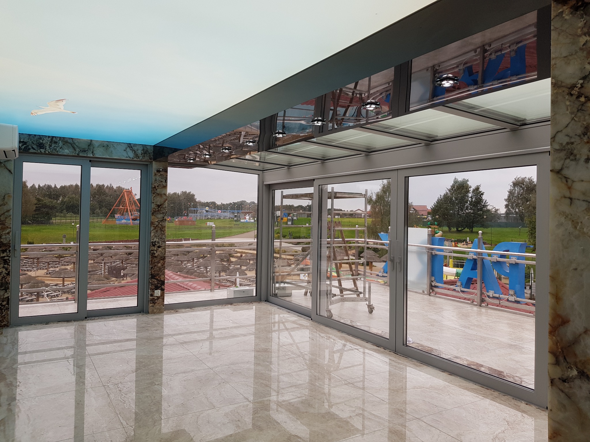 Wnętrze z przeszklonymi ścianami i sufitami, widok na zewnątrz na teren rekreacyjny z parasolami i konstrukcjami, marmurowe wykończenie filarów i podłogi, sufit podwieszany z motywem nieba i mewy.