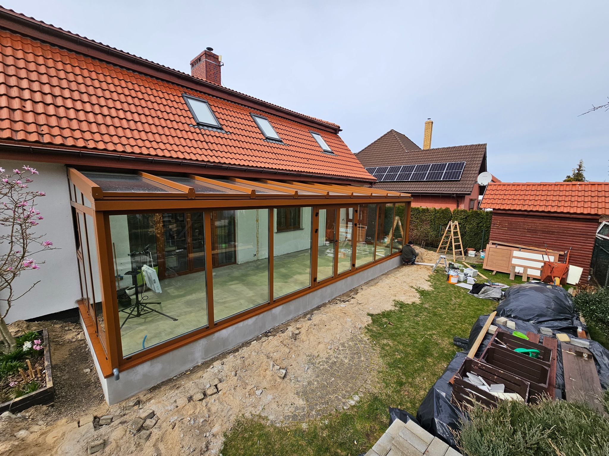 Zewnętrzny widok przeszklonej oranżerii z brązowymi ramami, przylegającej do domu z czerwoną dachówką, widoczny teren wokół w trakcie prac wykończeniowych.